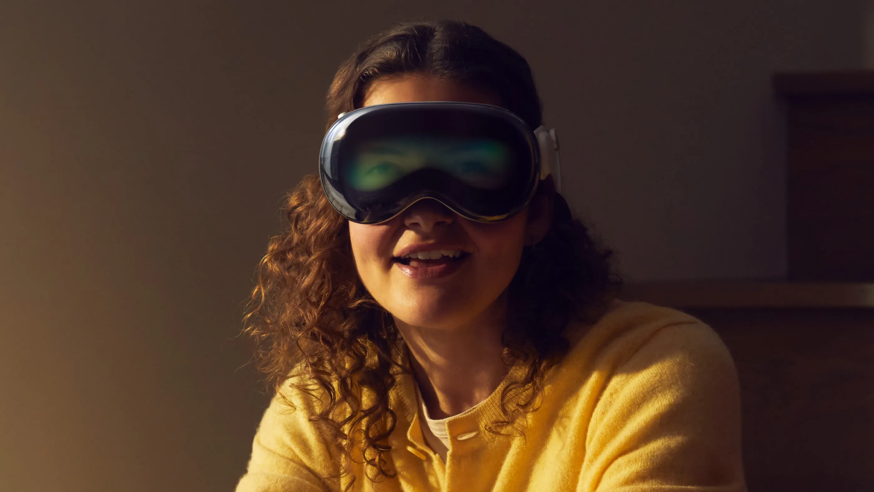A woman with long, curly, brown hair, wearing a yellow jumper is wearing Apple's Vision Pro virtual reality google.