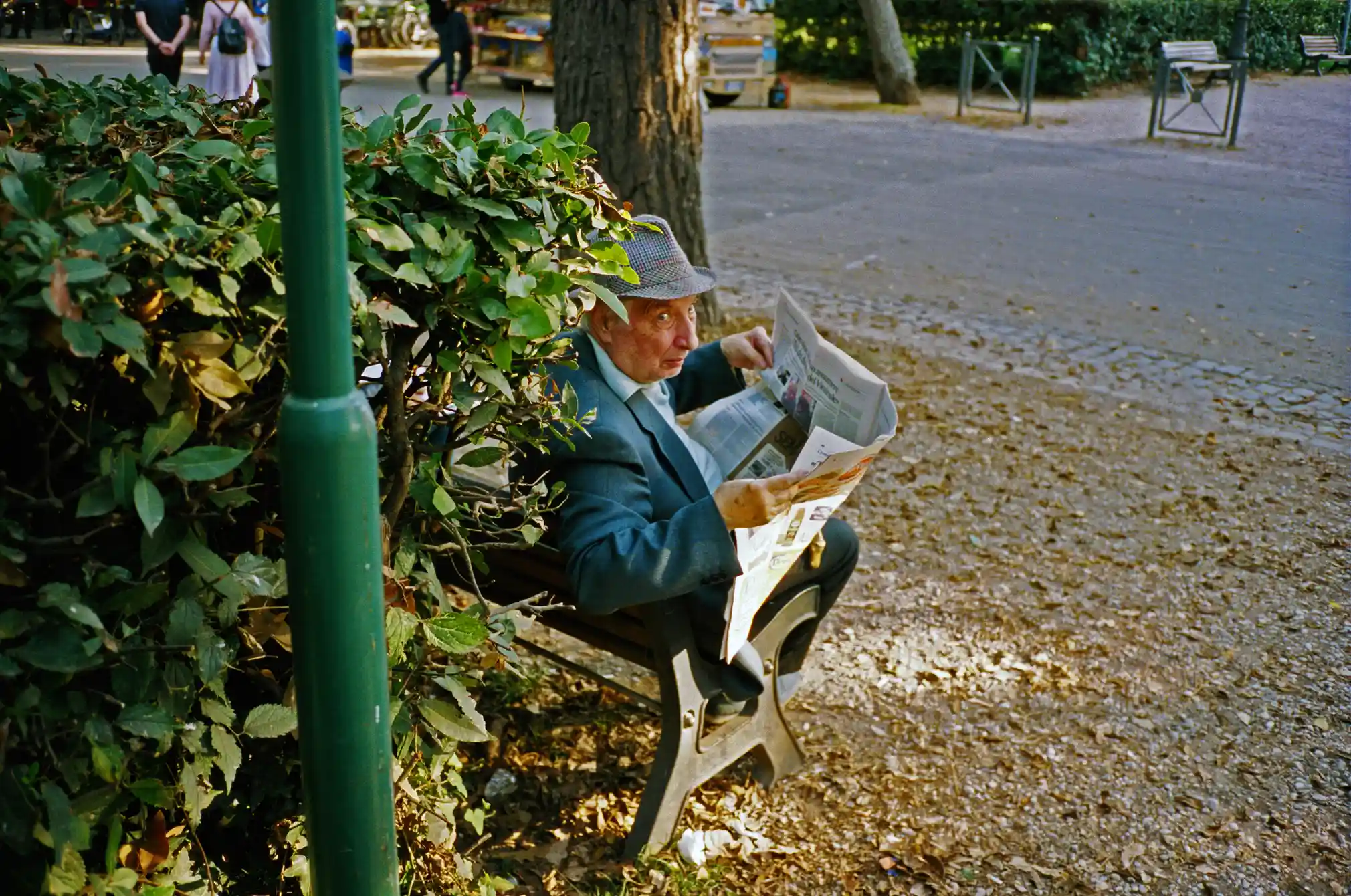 A man sitting on a bench with a newspaper. He is photographed from behind but peeks over his shoulder into the camera.