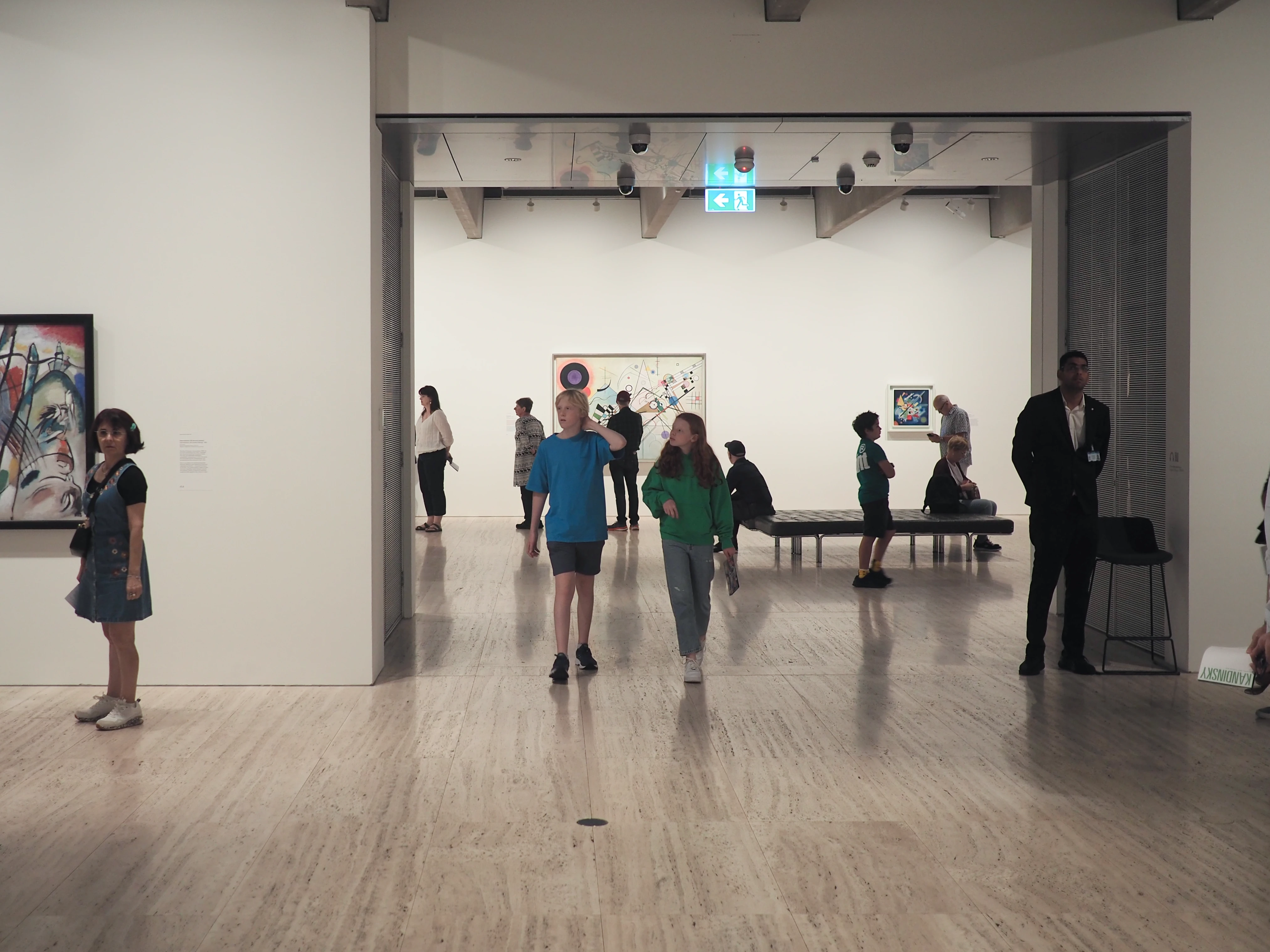 People walking through a bright-lit room in an art gallery, with Kandinsky art works on the walls.